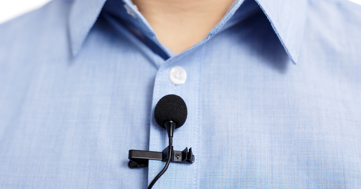 A black lavalier microphone with a clip and cord is fastened to the front of a light blue button-down shirt.