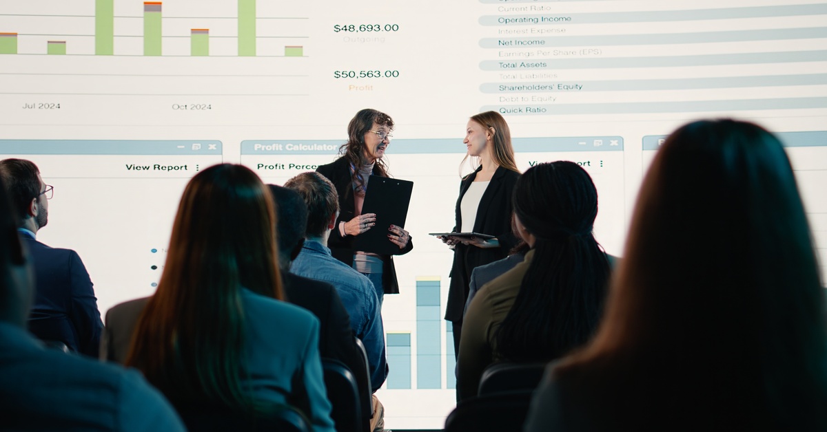 Two women stand before a seated audience with a microphone and folders as financial charts appear on a wall.