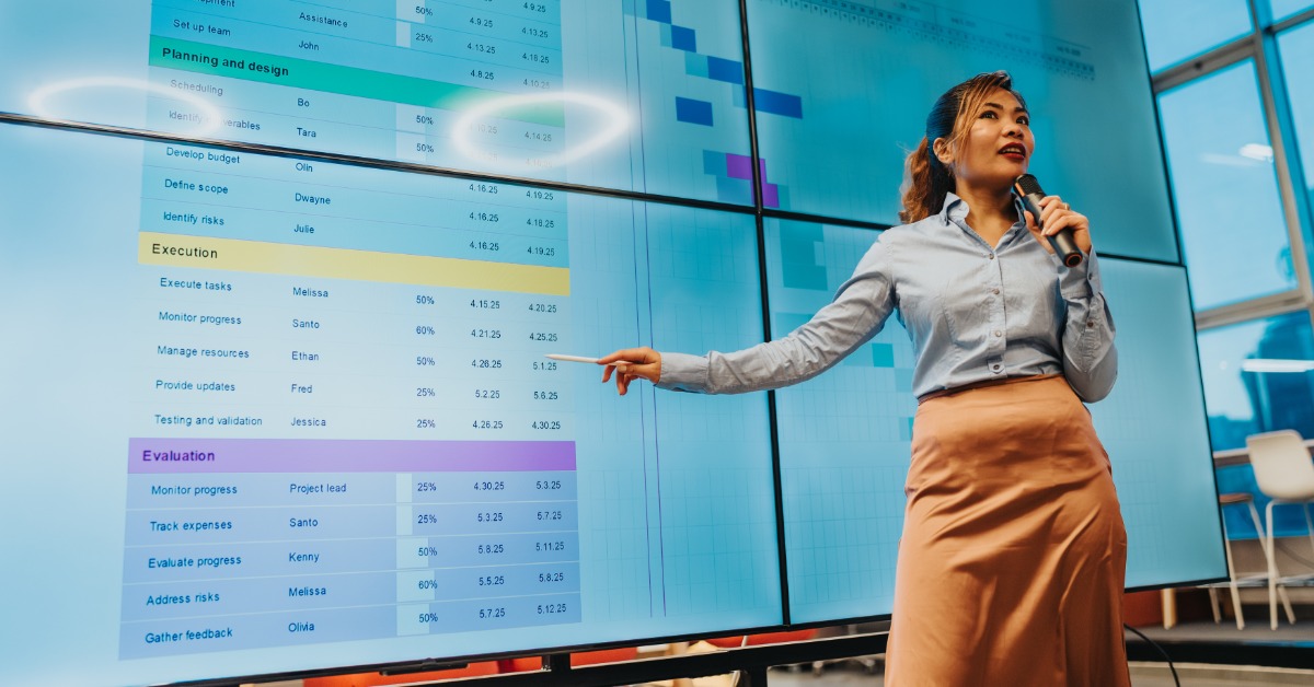 A woman holding a microphone points at large screens with charts and timelines. An overhead light reflects onto the screen.