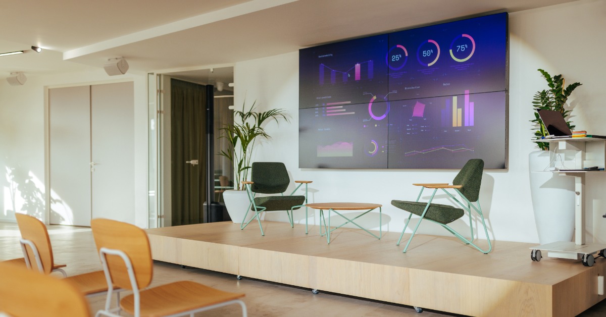 A conference room has chairs facing a small stage with two armchairs, a table, plants, and chart screens.
