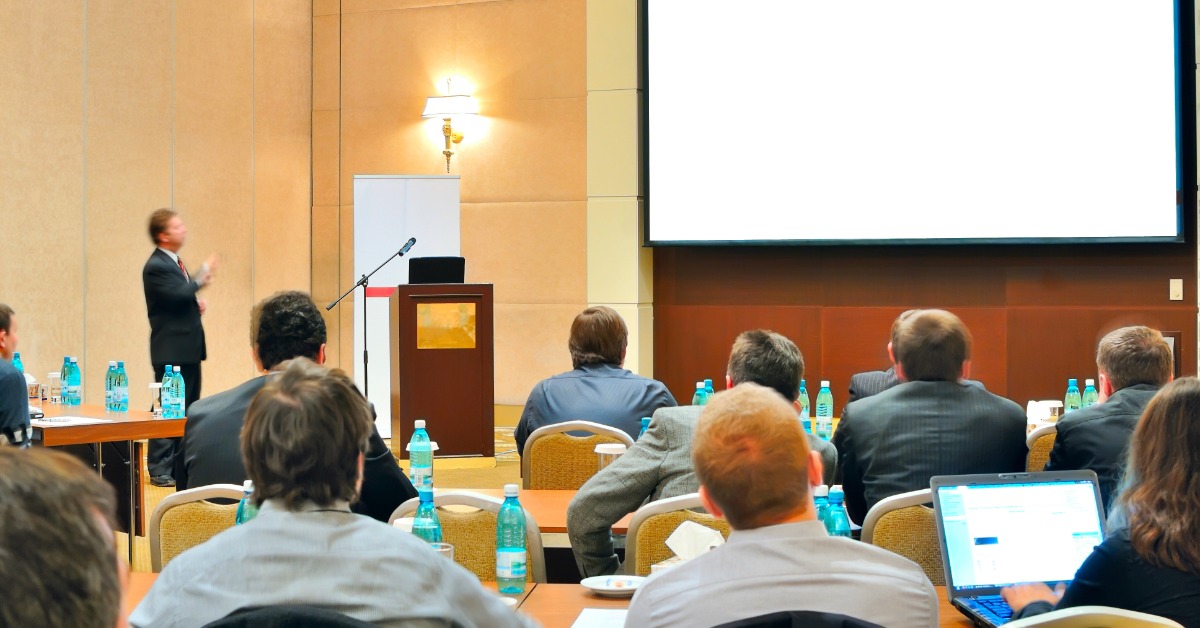 People seated at tables watch a presenter beside a podium and blank projection screen in a conference room.