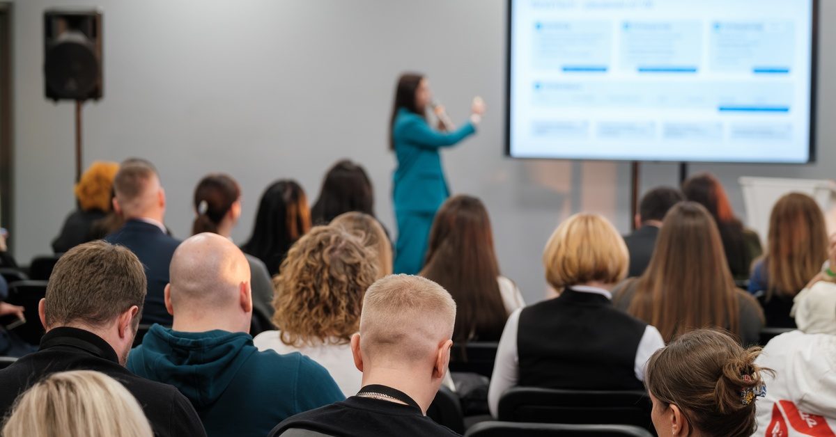 A seated audience faces a screen depicting charts. A person standing at the front of the room speaks into a microphone.